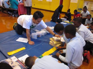 Children learning and doing experiments together on blue mats on the floor of a gym.